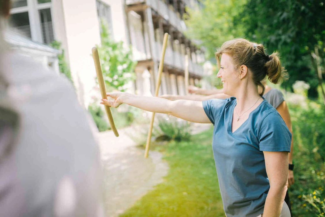 Frau macht l&auml;chelend &uuml;bungen drau&szlig;en in einer gruppe mit holz st&ouml;cken